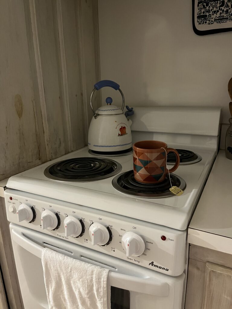 On a small white stovetop sits a Winnie the Pooh kettle on the back left burner and an orange mug with a red, green, orange, and white triangle pattern, a tea bag string visible on the rim, sits on the front right burner. There is a white towel hanging on the handle of the small oven below.