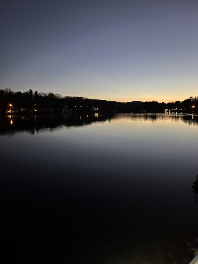 A scenic view of a lake at twilight, the orange to dark blue sky reflecting on the surface of the water. Some house lights are on along the shadow of a black tree line.