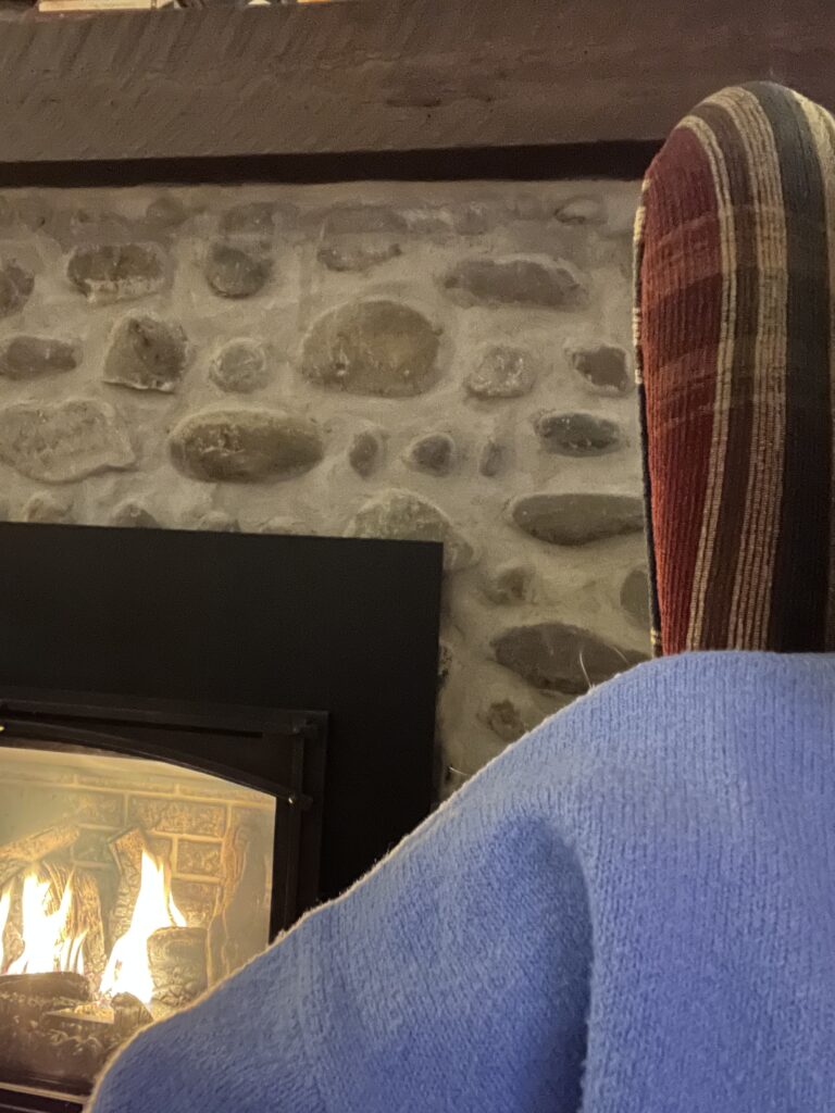 A stone fireplace sits in the background, a fire roaring inside. In the foreground, part of a plaid patterned wingback chair and the shoulder of a light blue sweater are just in frame.